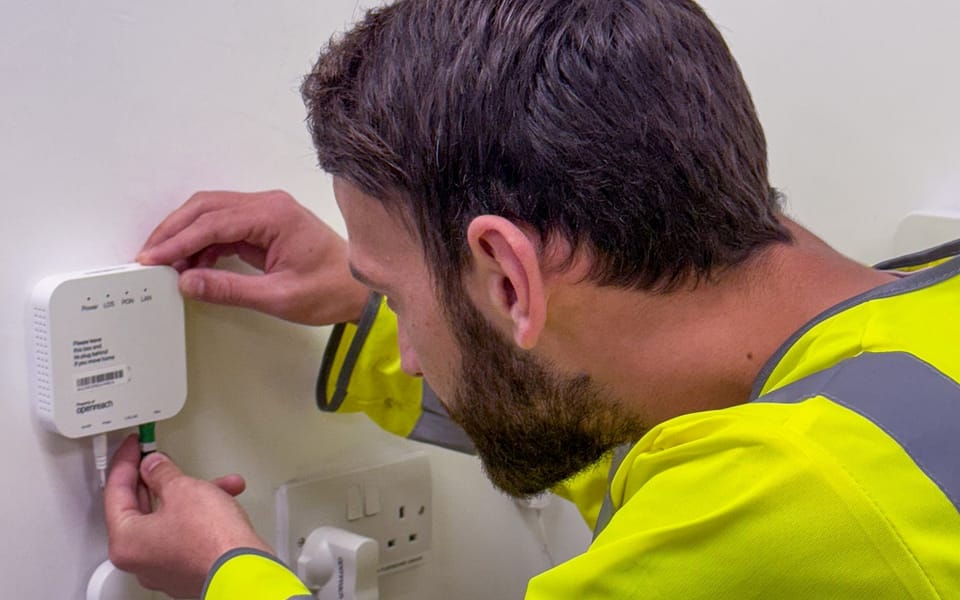 Openreach engineer in a high-visibility jacket fitting a fiber cable to an optical network terminal on a wall