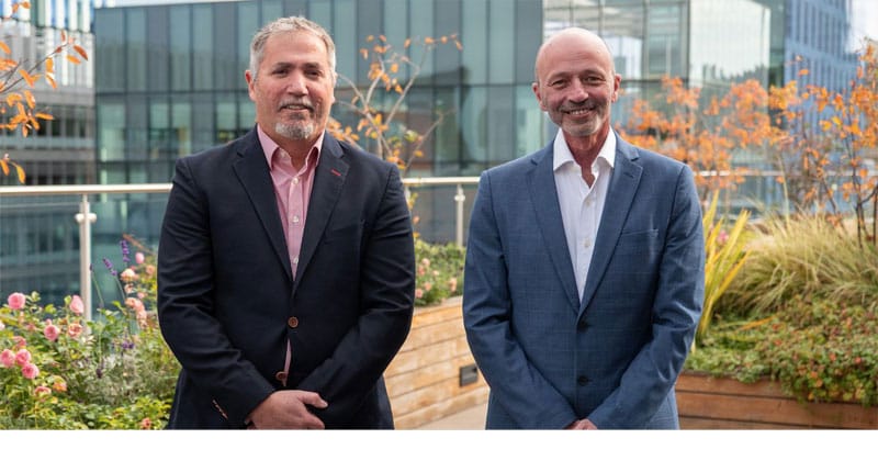 Two men in suits standing on a rooftop terrace with office buildings behind them, PEAK:AIO's photo for its £5m funding announcement