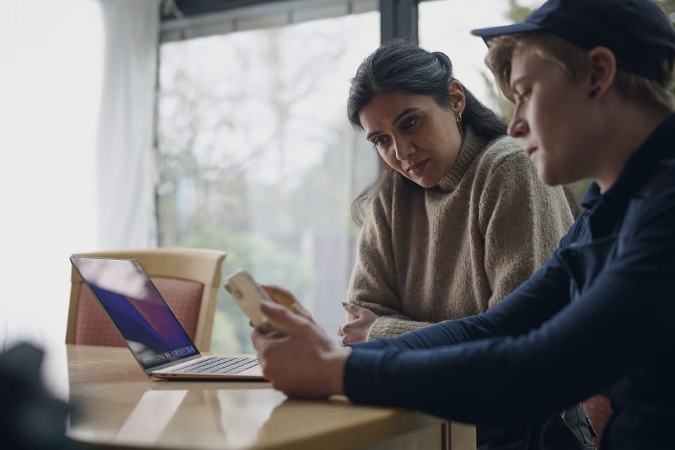 An Openreach engineer in a navy uniform and cap shows a phone screen to a woman sitting beside a laptop at a table by a window