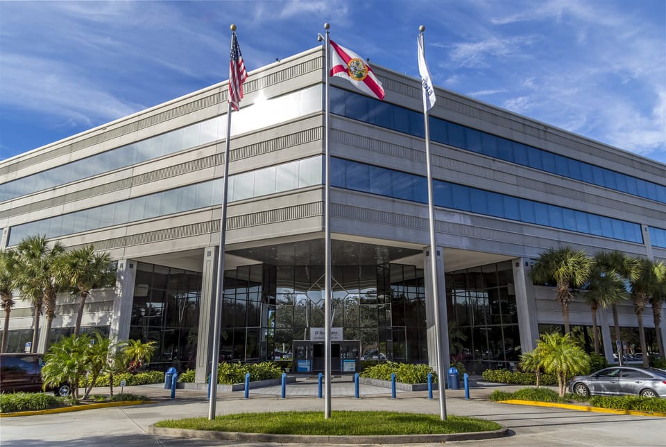 Exterior of a four-storey office building with glass window bands, palm trees and three flagpoles flying the US flag, the Florida state flag and a company flag, under a blue sky